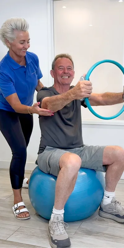 Nordic Balance practitioner guiding an older patient through a seated rehabilitation exercise using a resistance ring and stability ball.