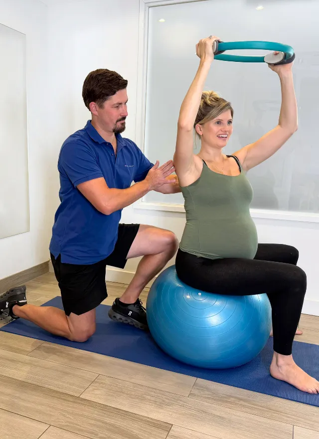 Pregnant woman performing a seated resistance exercise on a stability ball with guidance from a physiotherapist.