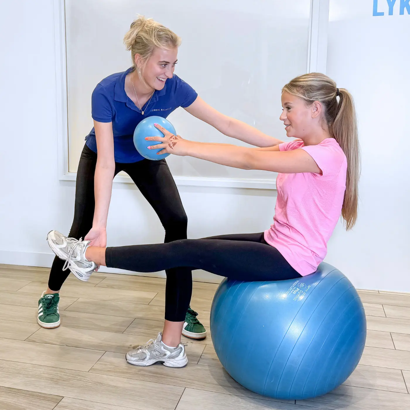 Paediatric osteopath at Nordic Balance guiding a teenager through a leg extension and balance exercise while seated on a stability ball.